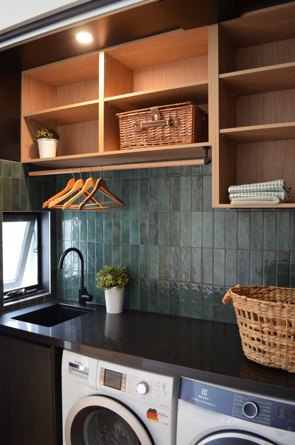 Modern laundry room with dark green tile backsplash, wooden open shelving, orange hangers, two washers below, and wicker storage baskets
