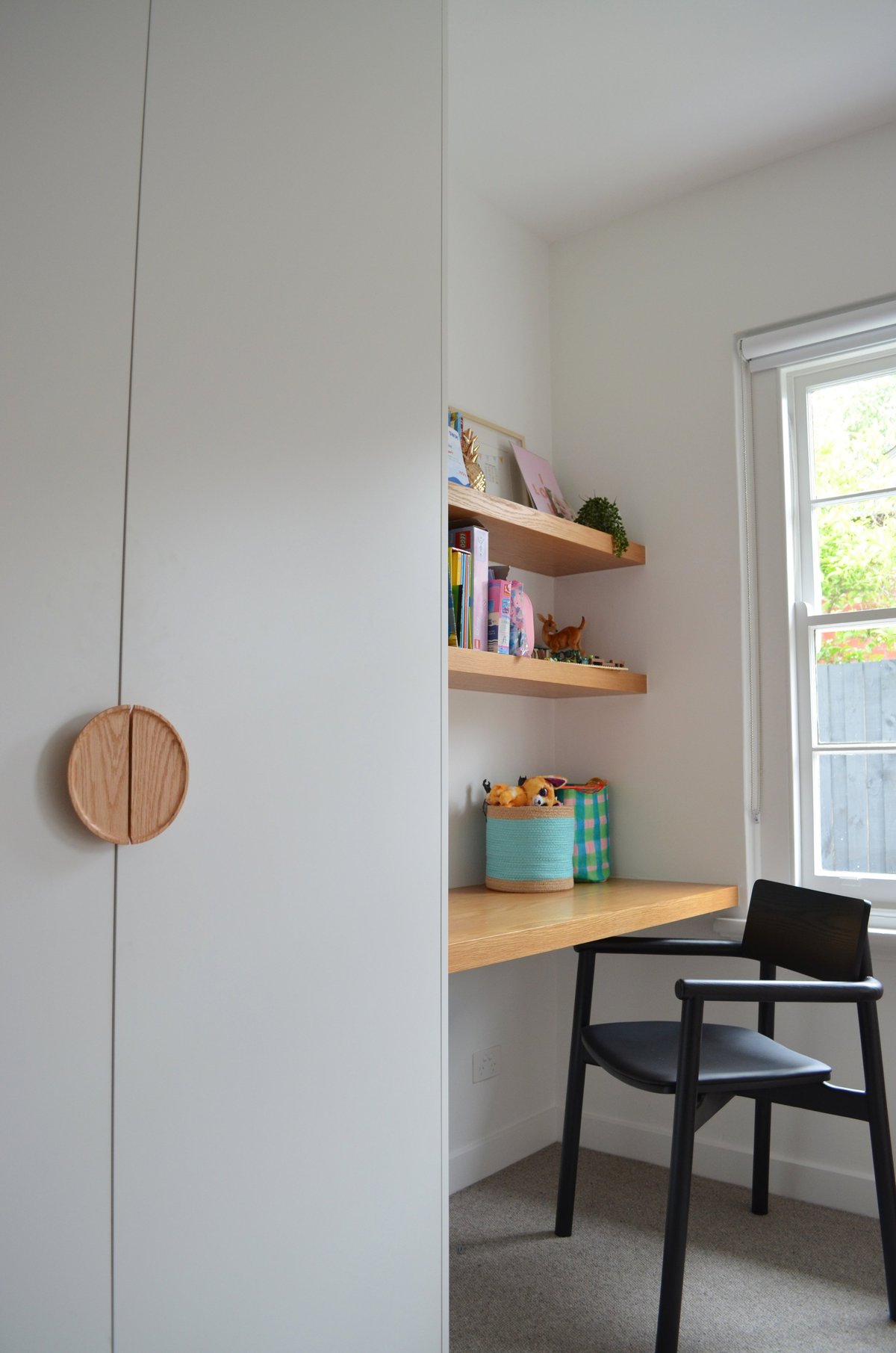 Modern home office nook with floating wooden shelves, wooden desk, black chair, and white built-in storage with circular handle