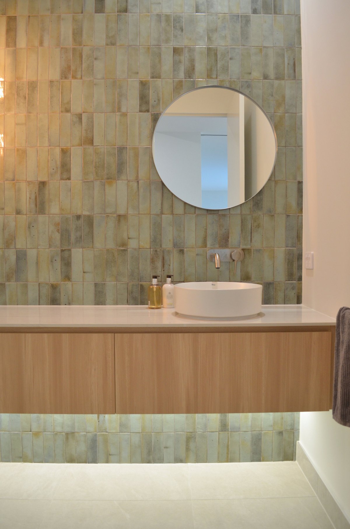 Modern bathroom vanity with round mirror, white vessel sink, and green tile backsplash above wooden cabinetry