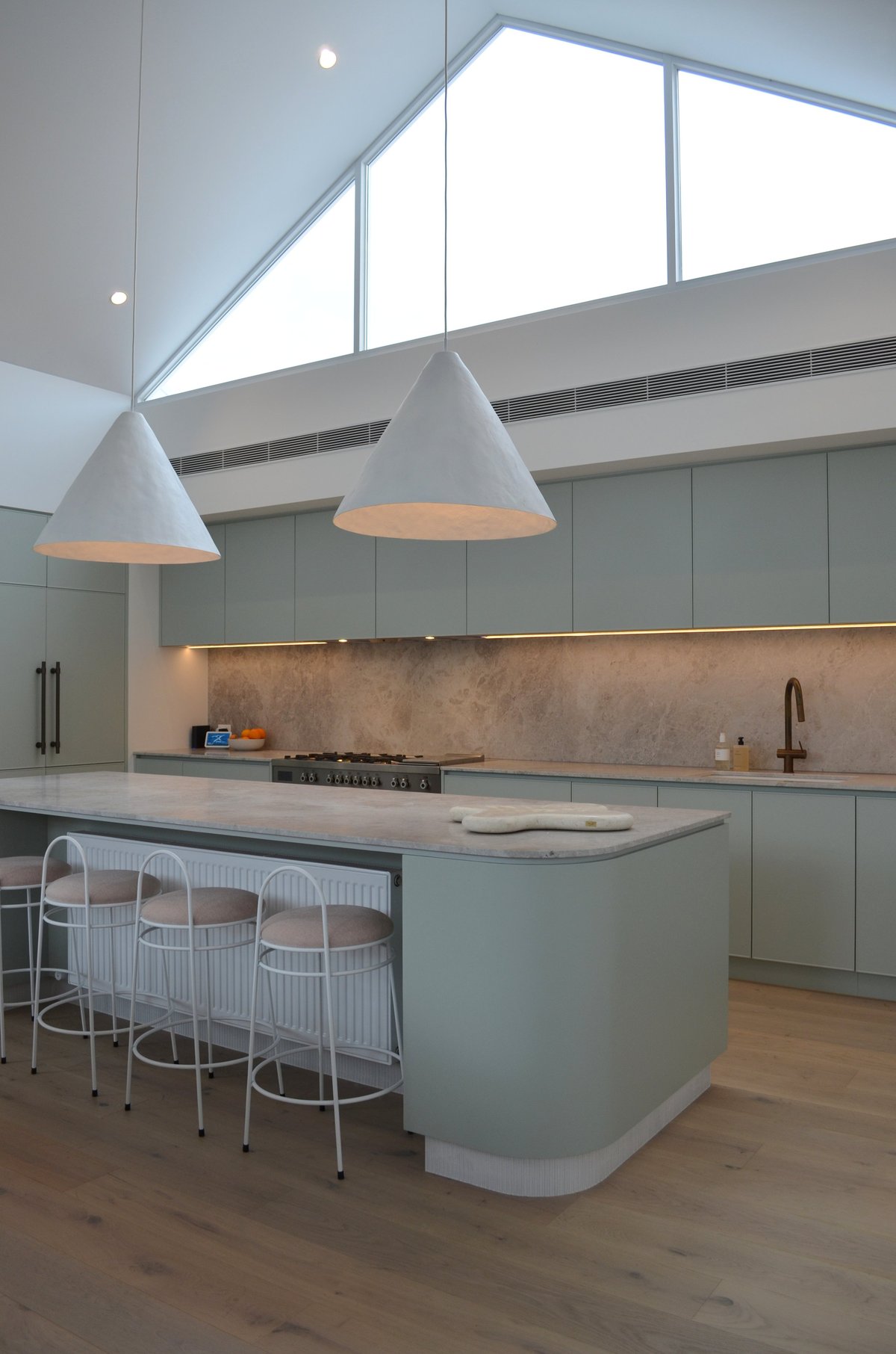 Modern kitchen with sage green cabinetry, concrete backsplash, gray cone pendant lights, island with bar seating, and skylights above.