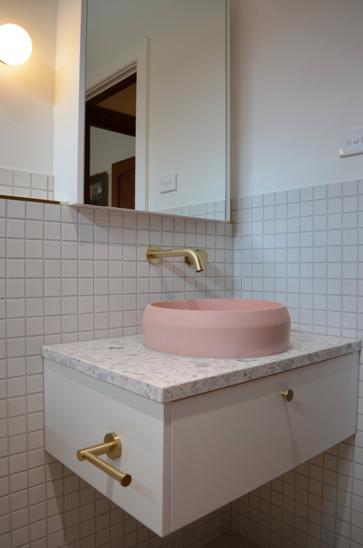 Modern bathroom with pink round vessel sink on white marble countertop, brass fixtures, wall-mounted mirror, and white tile walls