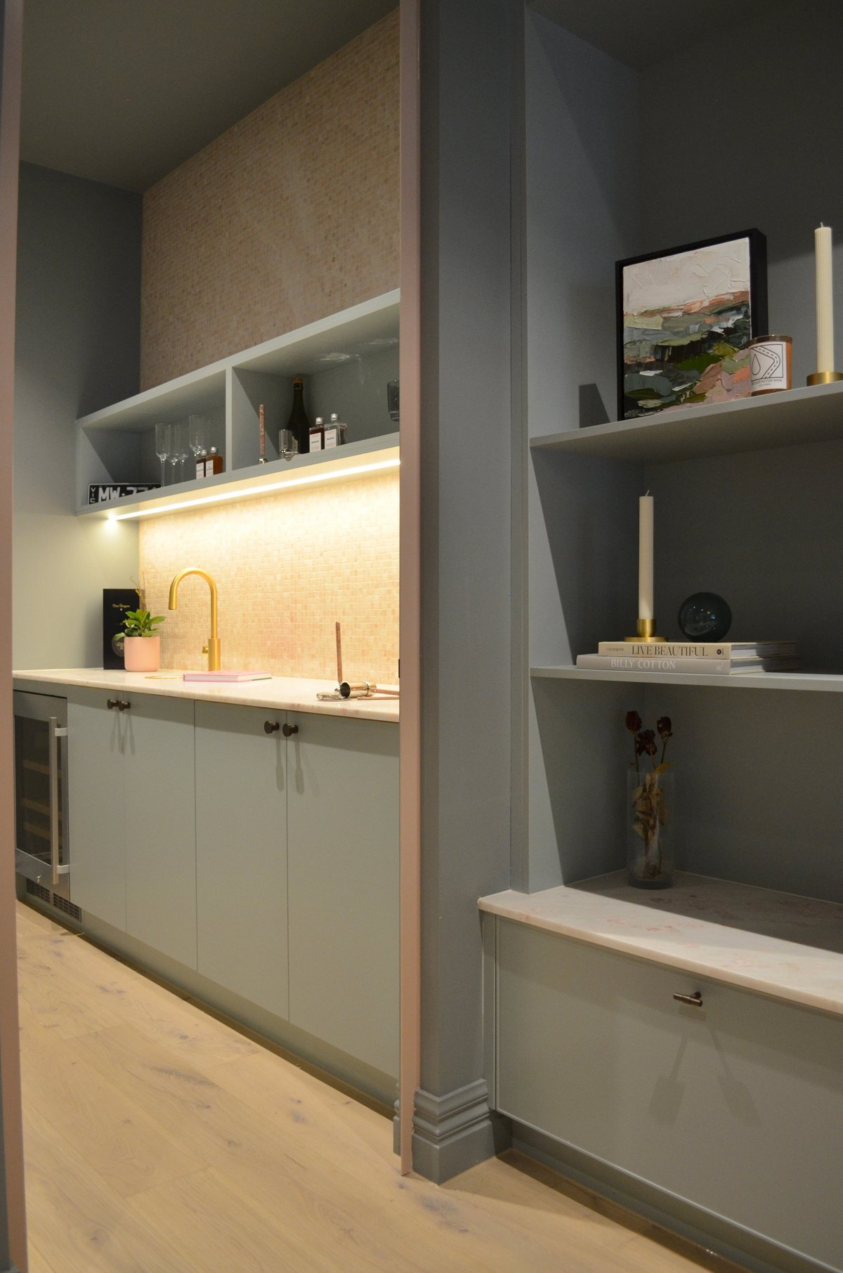 Modern kitchen with white cabinetry, warm wood accents, illuminated backsplash, and shelving unit with books and decorative items