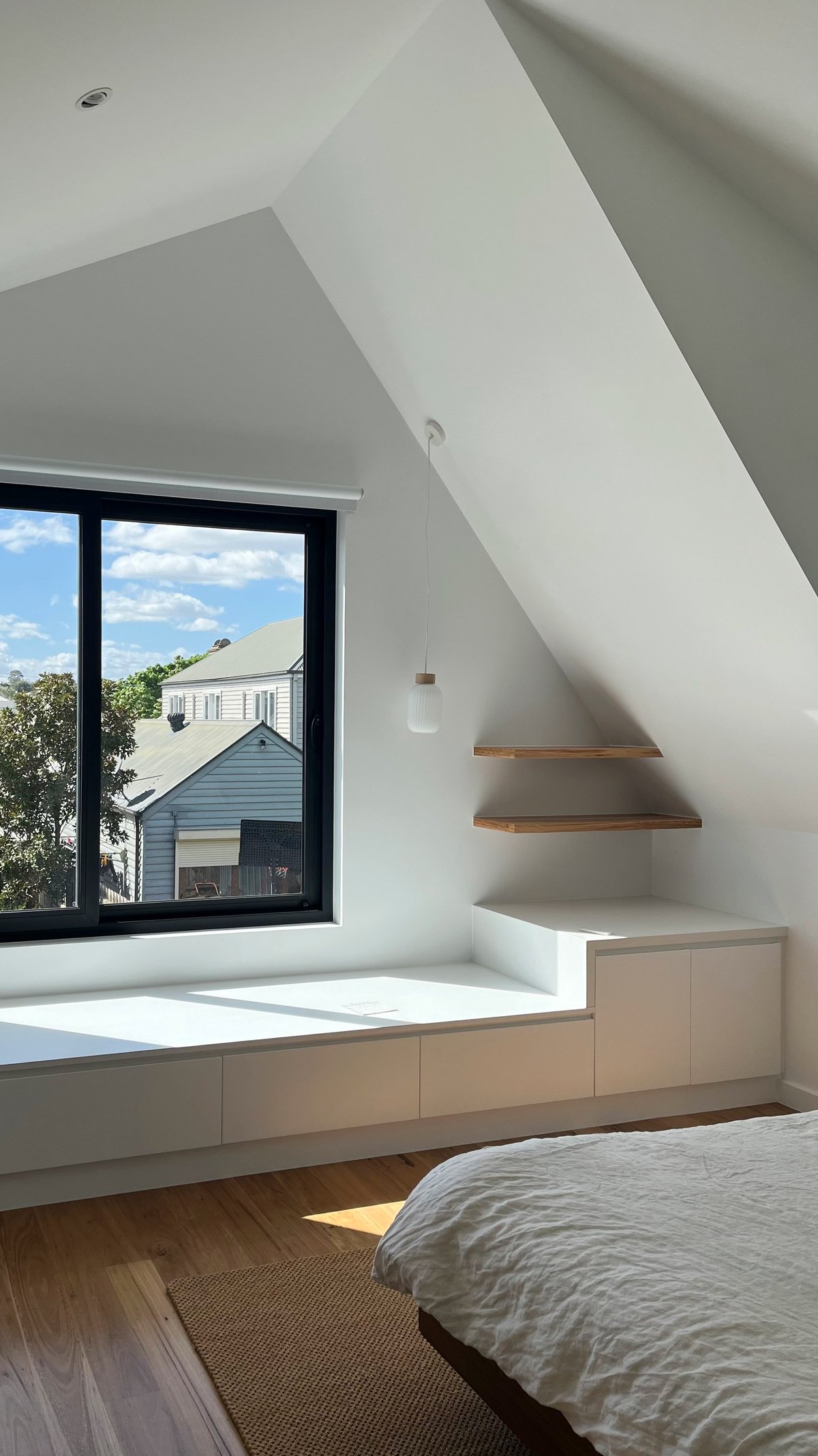 Modern bedroom with sloped ceiling, window seat with storage, wooden shelves, black framed window overlooking residential area, and hardwood flooring