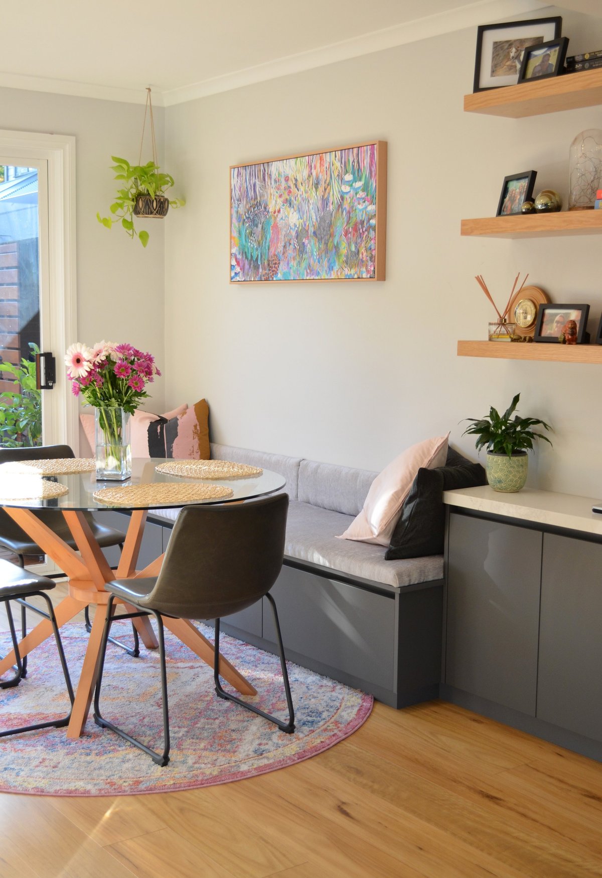 Bright modern living room with round dining table, black chairs, gray built-in bench, wooden shelves, colorful artwork, fresh flowers, and plants