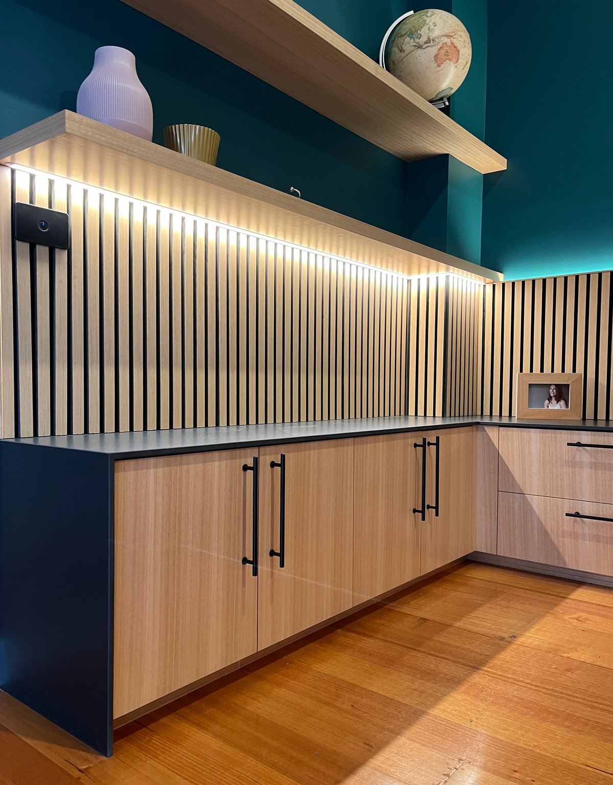 Modern kitchen with wooden cabinets, illuminated white slatted backsplash, teal wall, floating shelf, and wooden flooring.